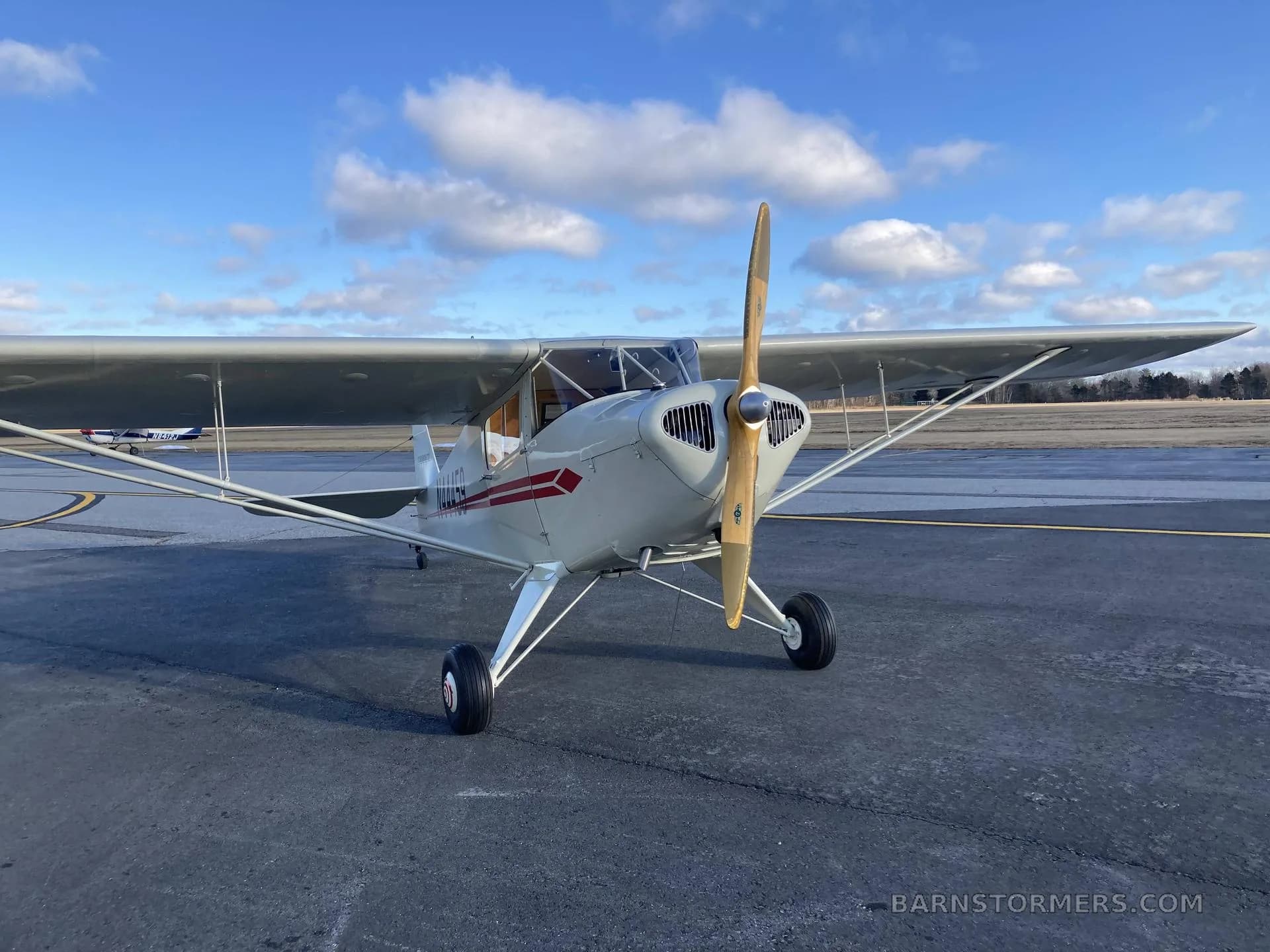A front view of a white and red Piper Cub N44459 with a wooden propeller, parked on an airfield under a partly cloudy sky.
