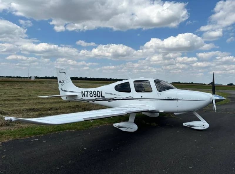 Side view of a white Cirrus SR22 aircraft with black accents and registration N789DL on a grassy airfield.