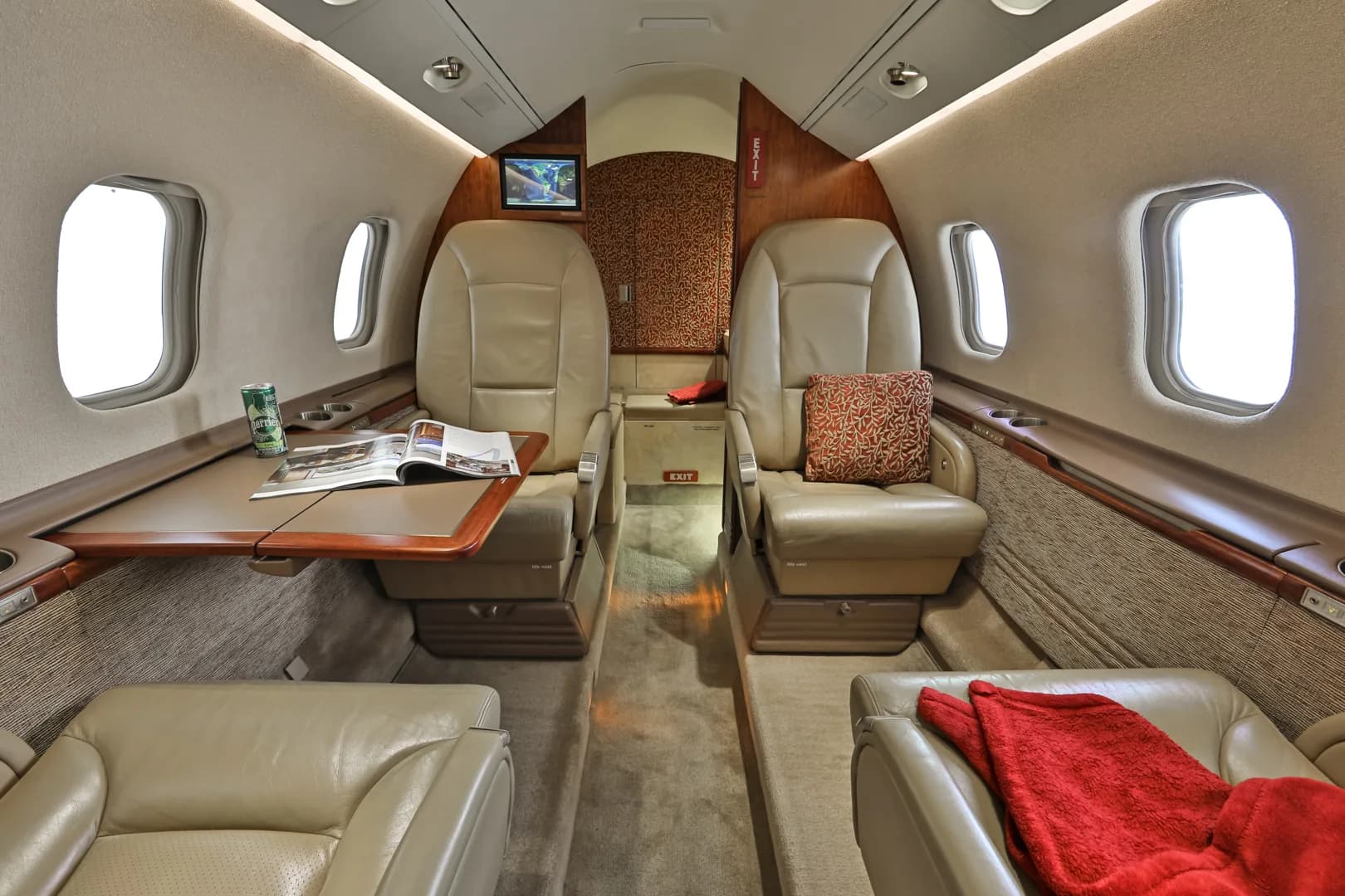 The spacious interior cabin of a business jet, showing beige leather seating, a fold-out table, and a small monitor at the front.