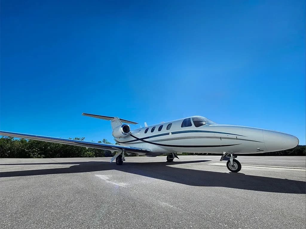 A white private jet with blue and grey stripes sits on an asphalt tarmac under a clear blue sky.