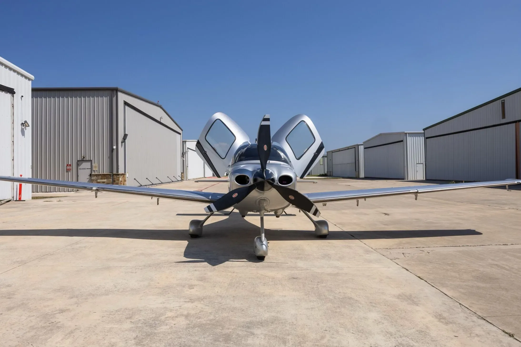A head-on view of the silver and black Cirrus SR22 aircraft with its gull-wing doors open, parked on a sunny tarmac.