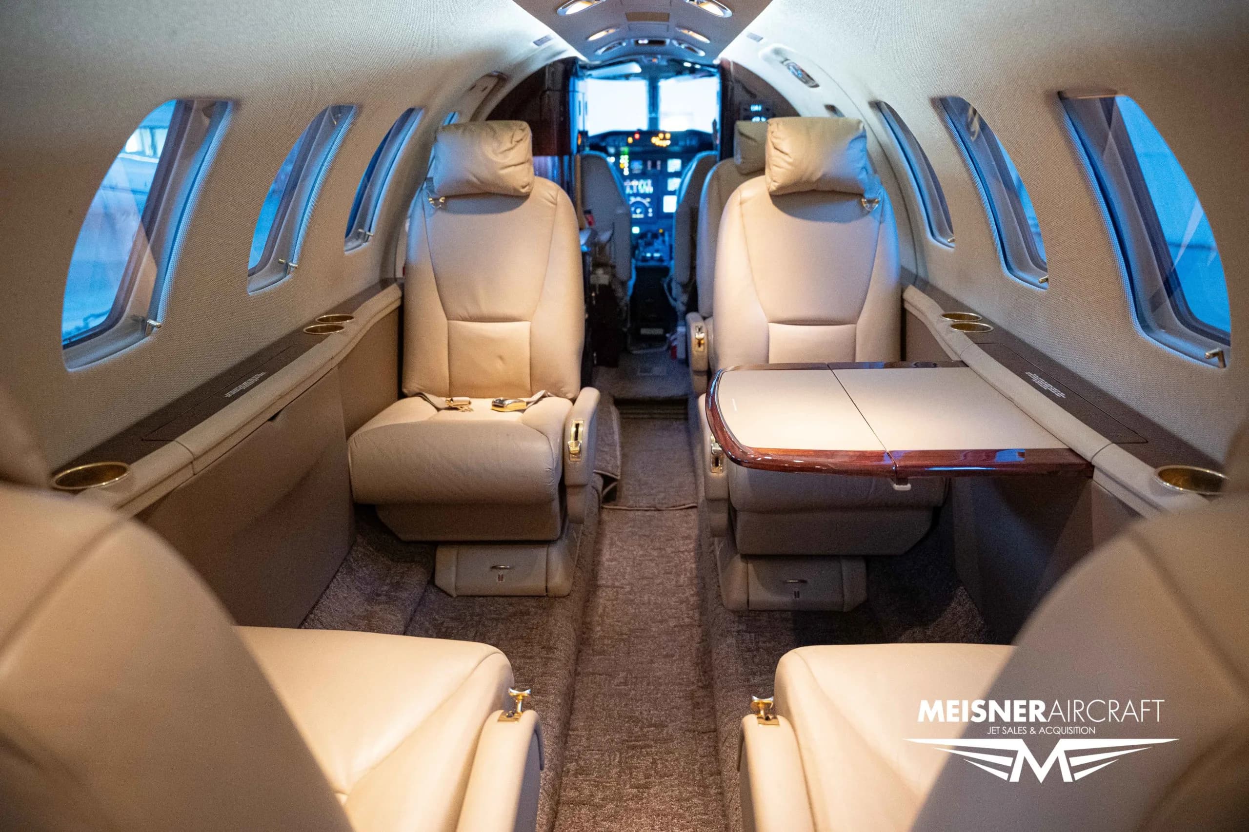 The interior cabin of a private jet, featuring beige leather seats and a wooden fold-out table, looking towards the cockpit.