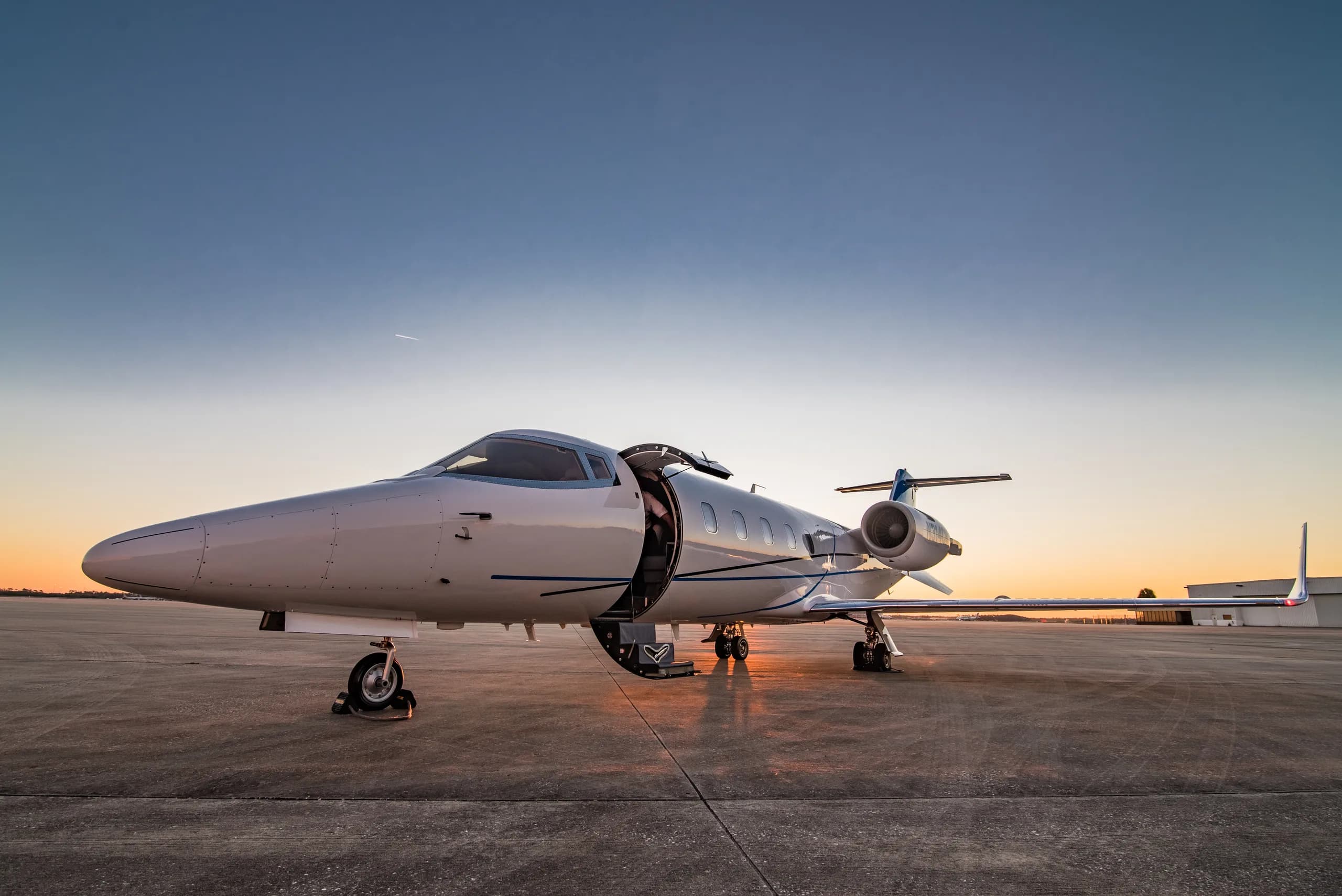 A white business jet with its main door open, parked on an airfield at sunset with a warm glow.