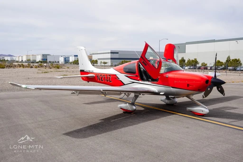 A red and silver Cirrus SR22 aircraft with open doors is parked on an asphalt ramp with hangars in the background.