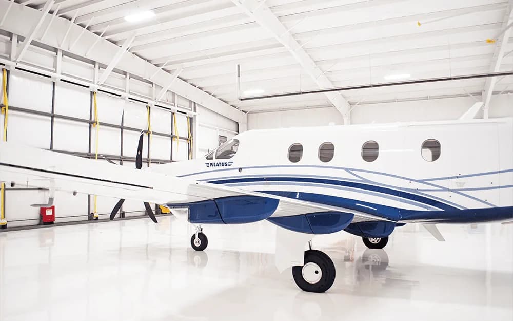 A side view of the white and blue Pilatus PC-12 aircraft, showing the landing gear and fuselage in a hangar.