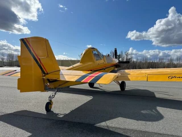 Rear three-quarter view of a yellow crop duster aircraft showing its tail, wing, and landing gear on an airfield.