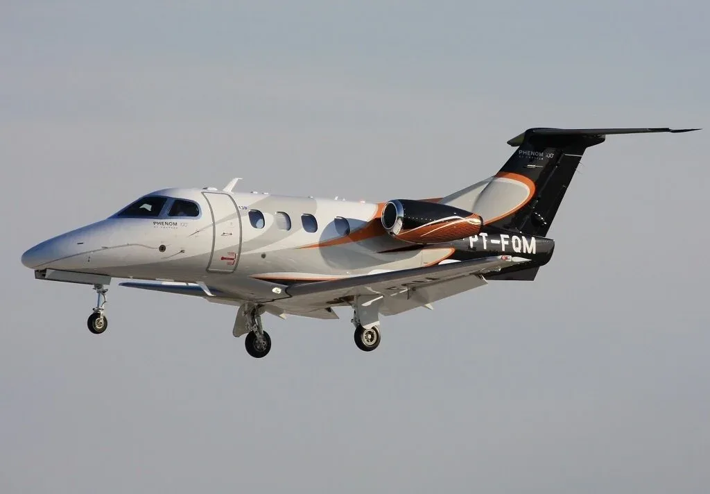 A Phenom 100 aircraft with registration PT-FOM is shown in flight with a gray and orange paint scheme.