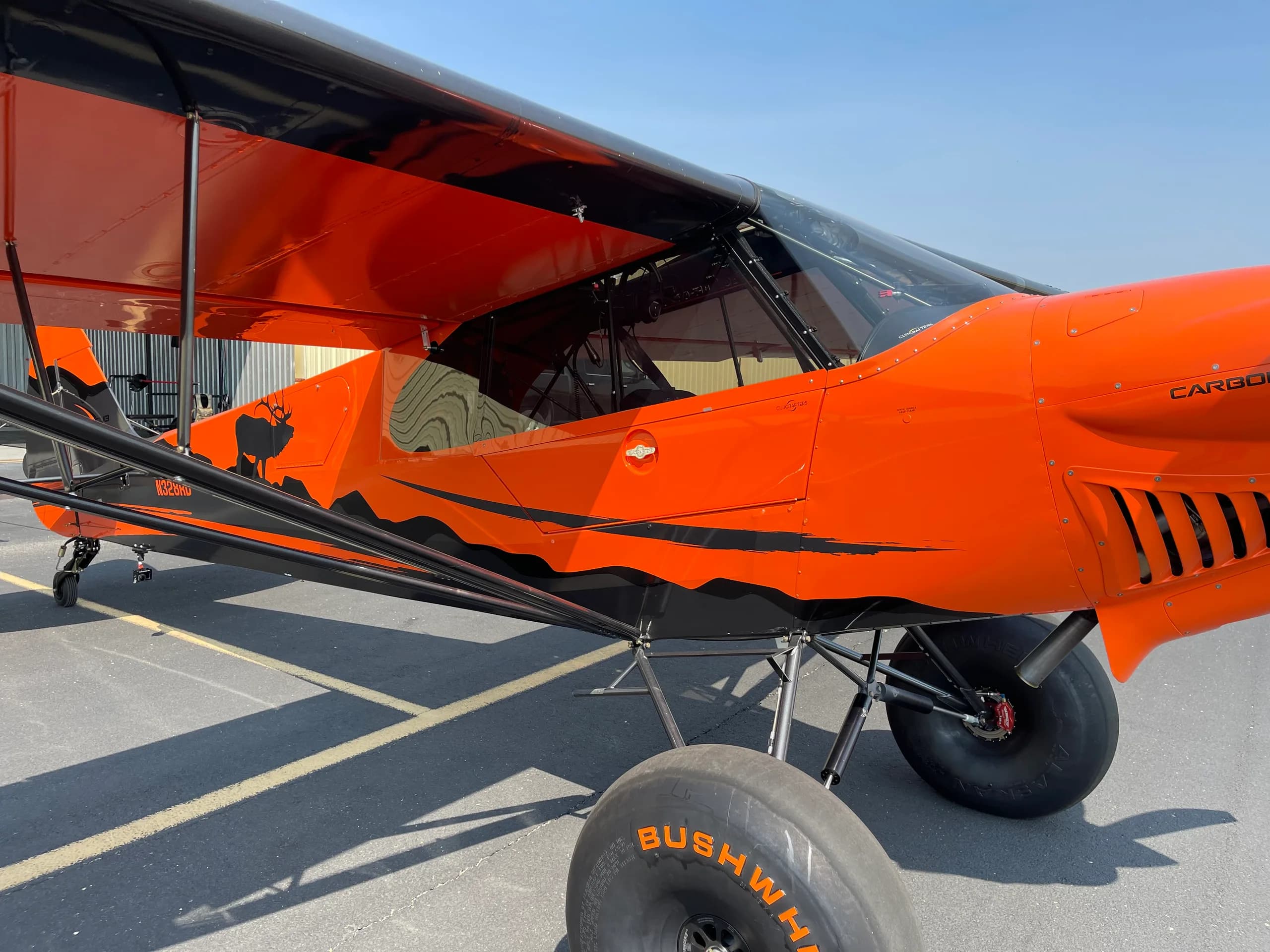 A close-up side view of the orange and black Carbon Cub FX-3 aircraft, showing the cockpit area and large bush wheels on an airport tarmac.