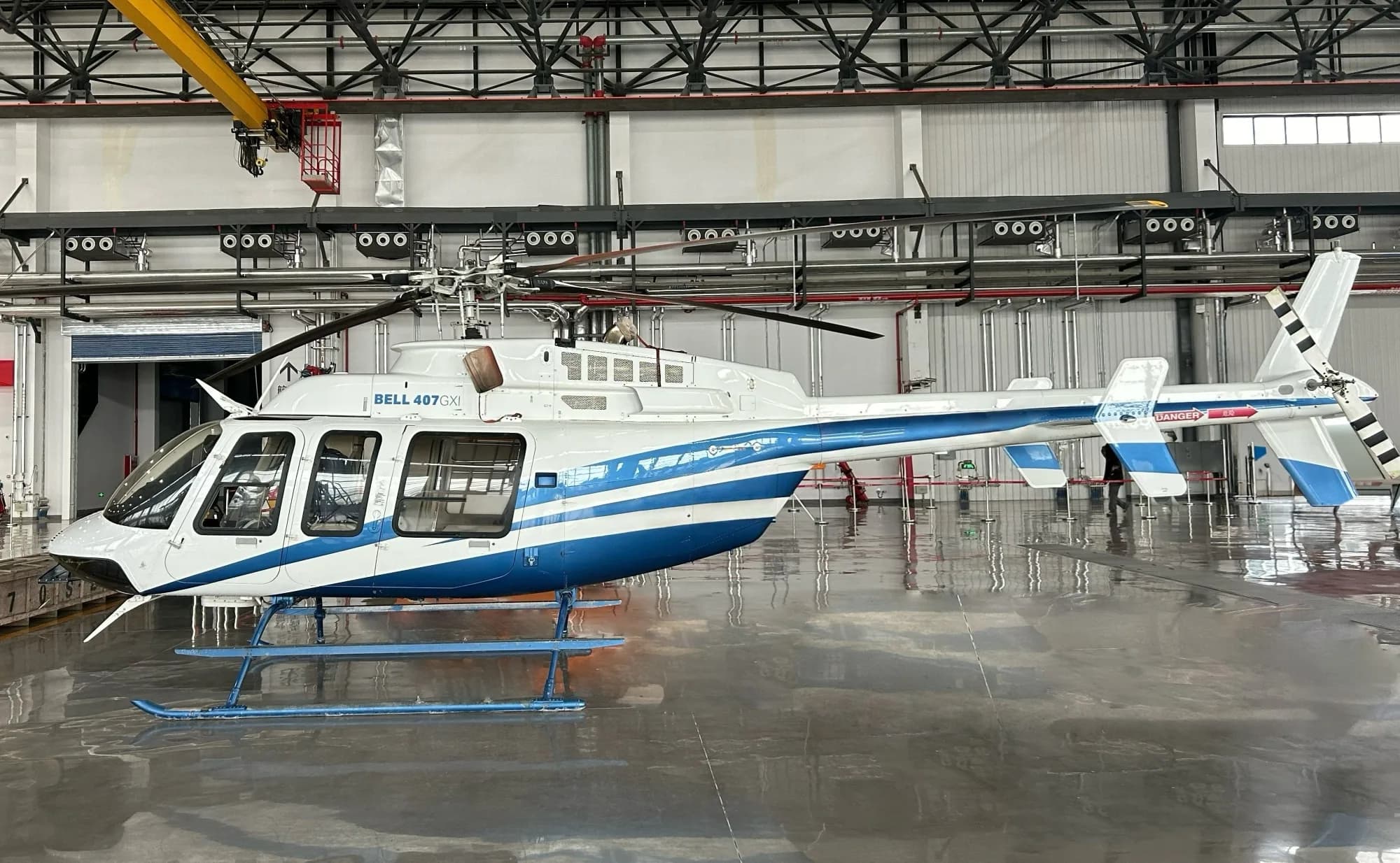 A white Bell 407GXI helicopter with blue stripes sits in a hangar with a reflective floor.