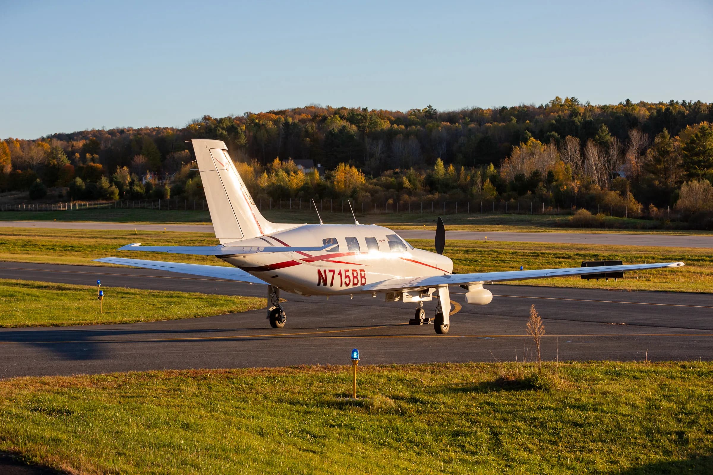 A three-quarter rear view of the Piper Malibu Mirage N715BB on a runway with autumn foliage.