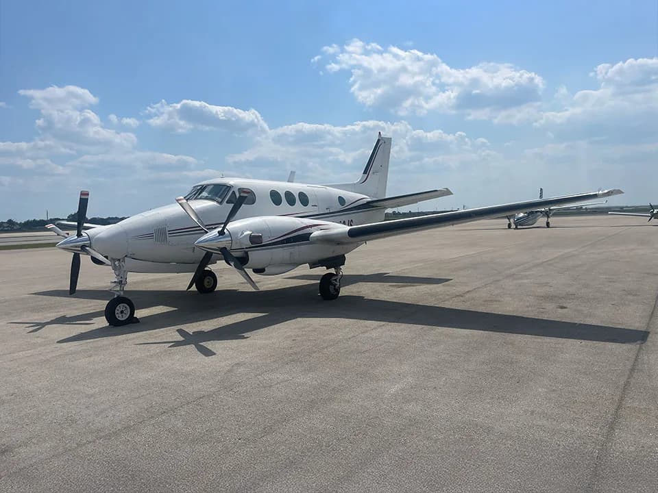 A white twin-engine turboprop aircraft with dark stripes on the wings and tail, parked on an airfield under a partly cloudy sky.