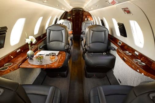 A wide view of a private jet's elegant cabin with black leather seating, a wooden table set with drinks, and bright windows.