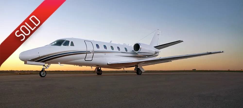 A white Cessna Citation business jet with brown and gold stripes is parked on an airfield at sunset.