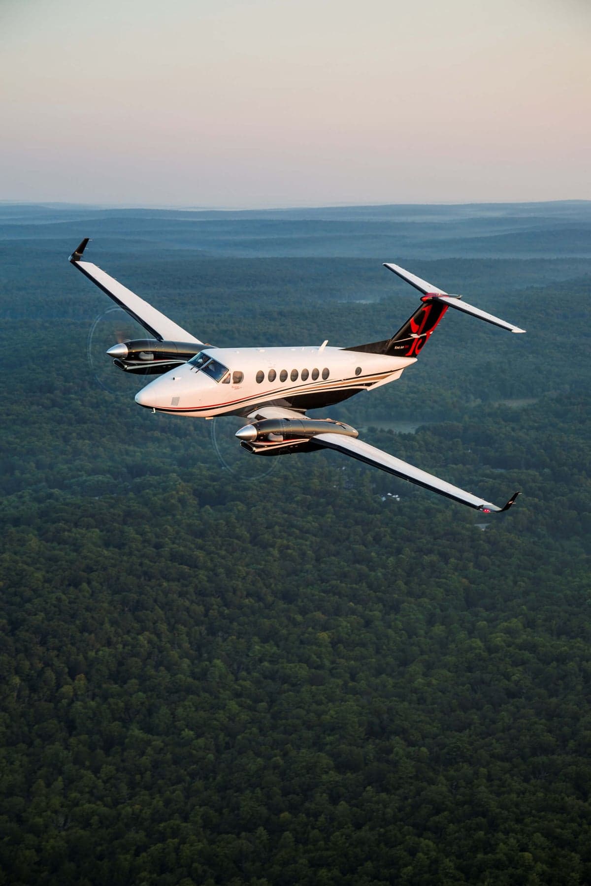 A white and black King Air 350i with registration N831KA flies over a dense green forest.