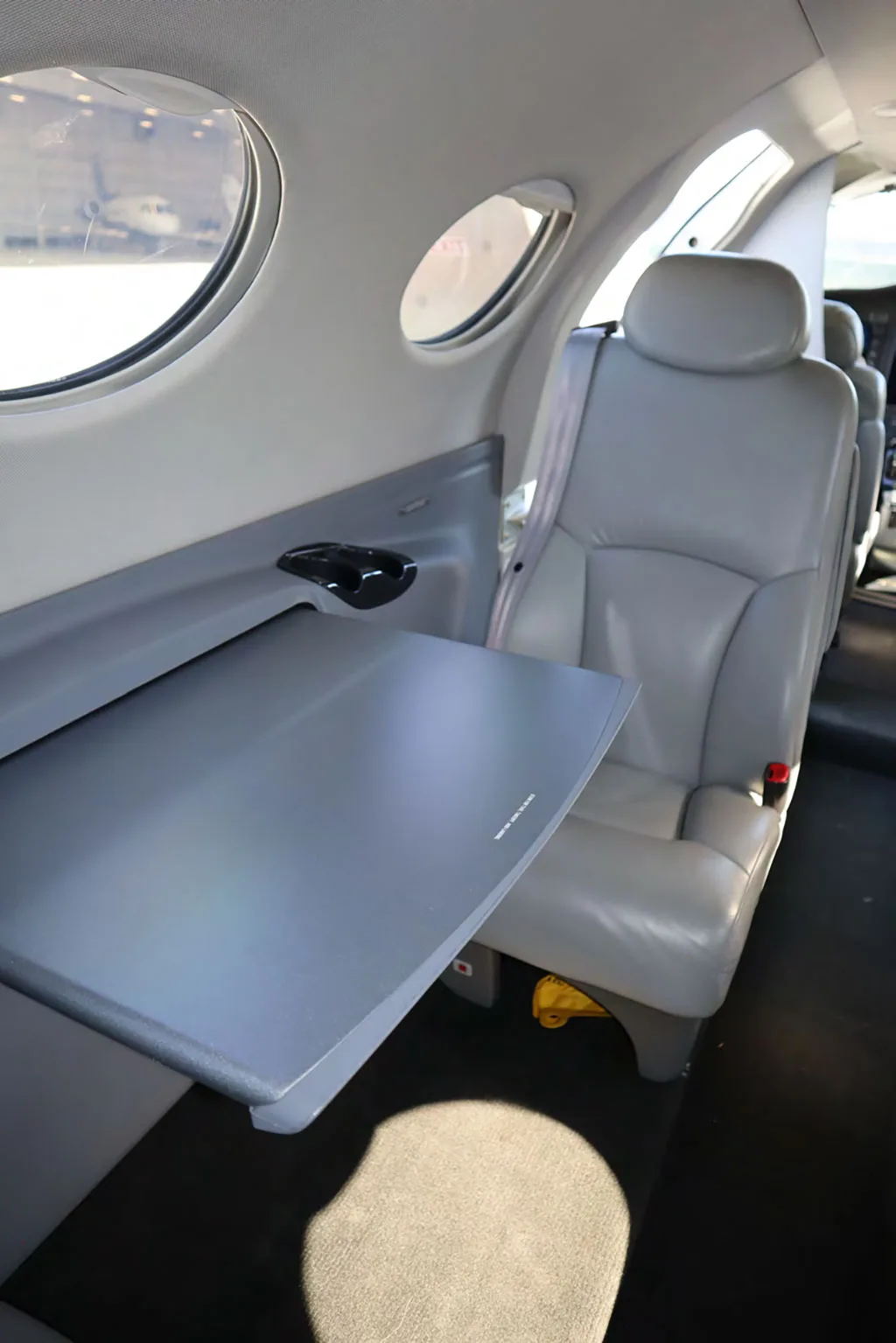 A close-up view of a light gray fold-out table next to a window and a seat in the aircraft cabin.