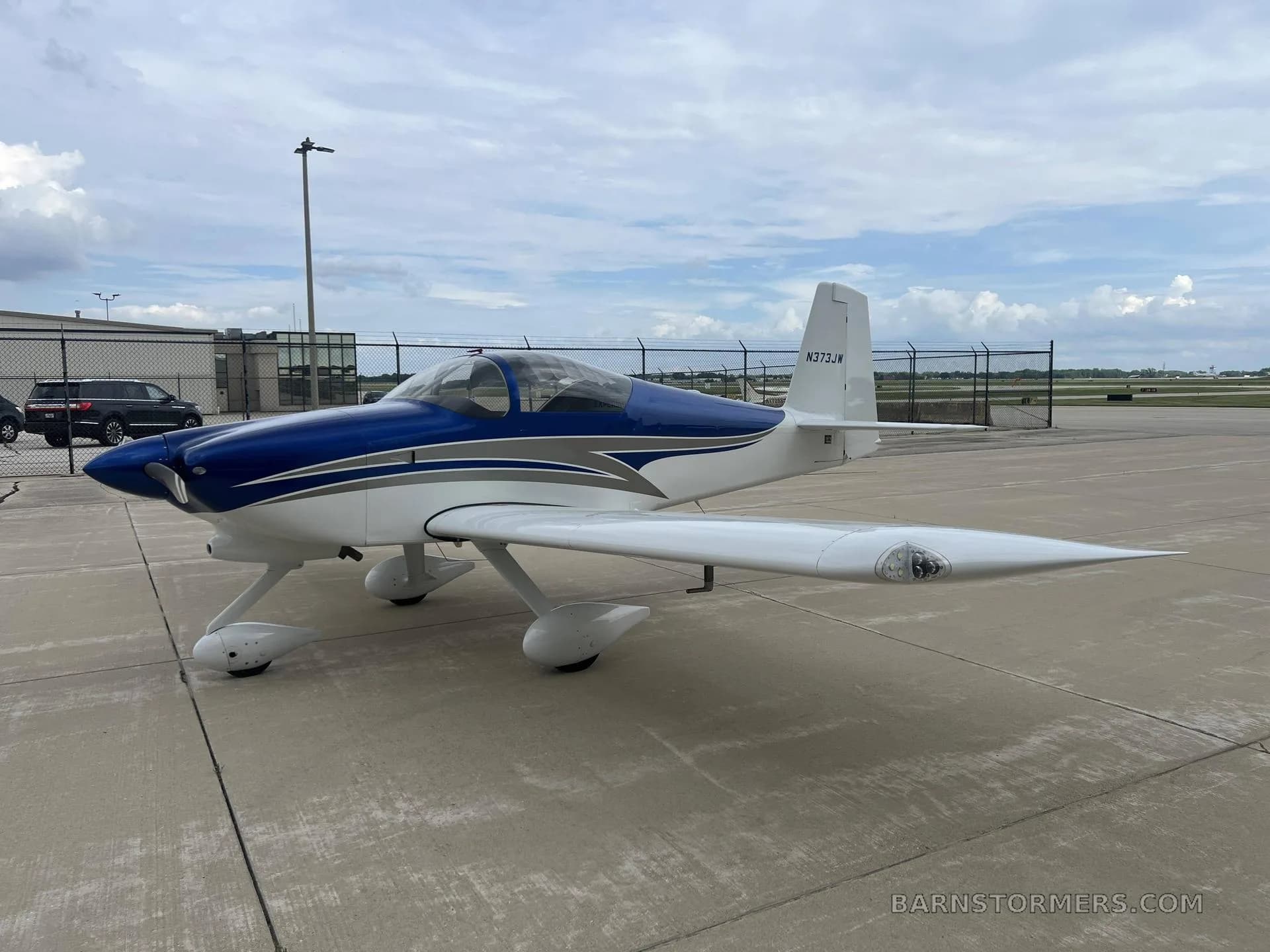 A white and blue single-engine aircraft, N373JW, with a bubble canopy, parked on a concrete tarmac under a cloudy sky.