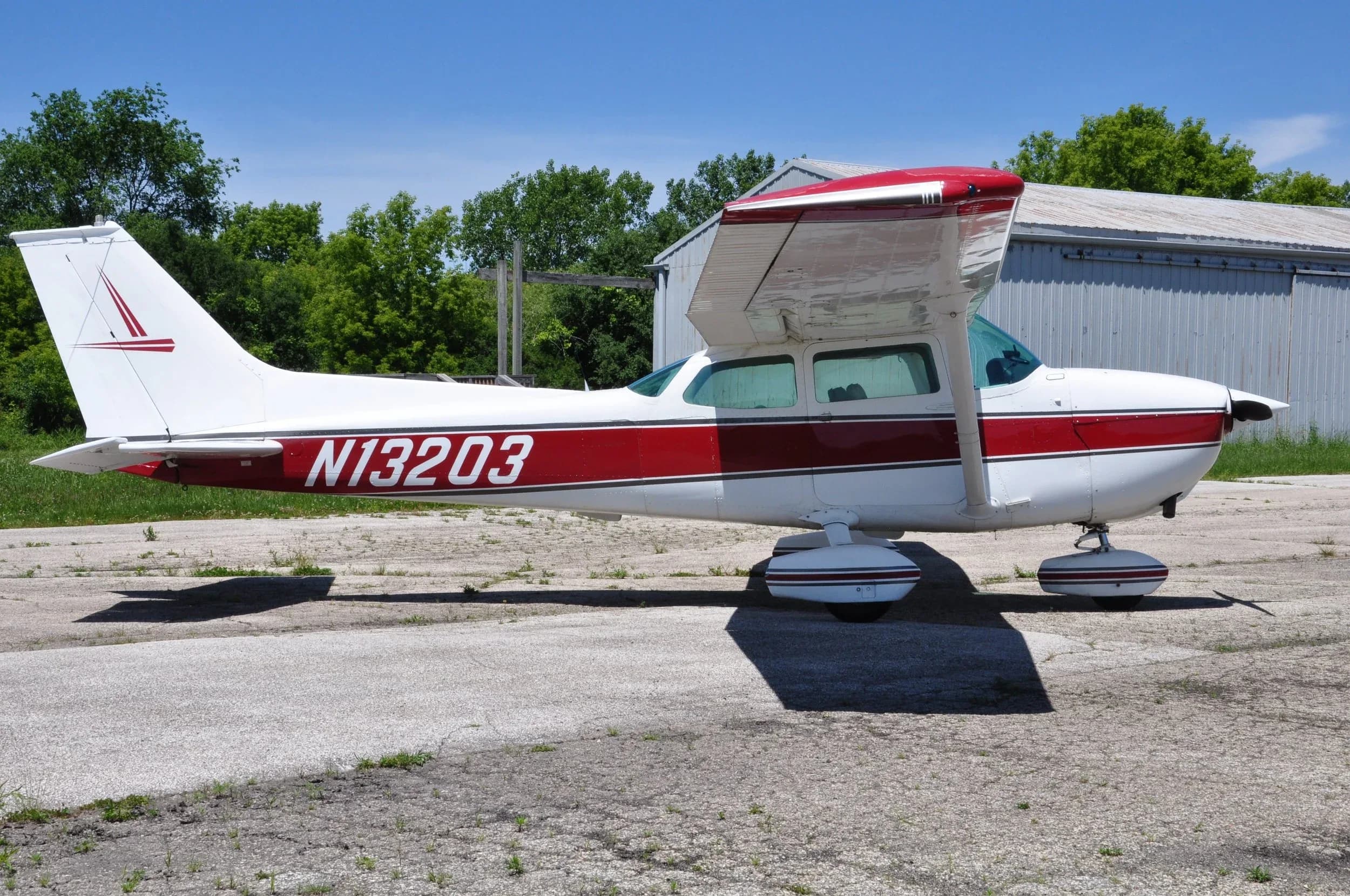 A side view of a white and red Cessna 172 aircraft (N13203) parked on an asphalt ramp next to a hangar.