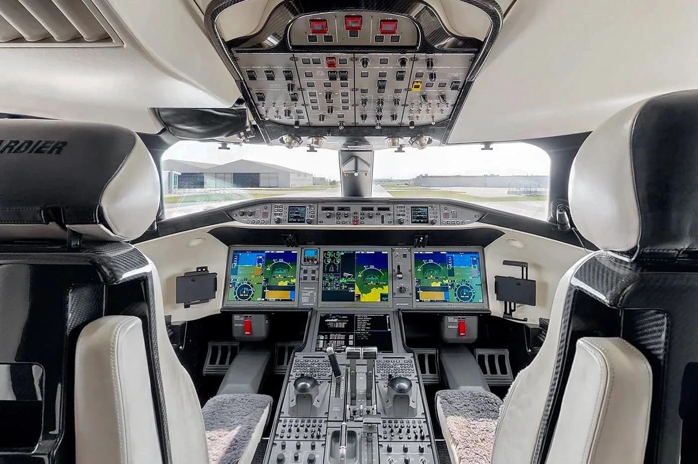 The modern cockpit of a private jet, featuring multiple digital displays, overhead panel, and control yokes, with a view of a hangar in the distance.