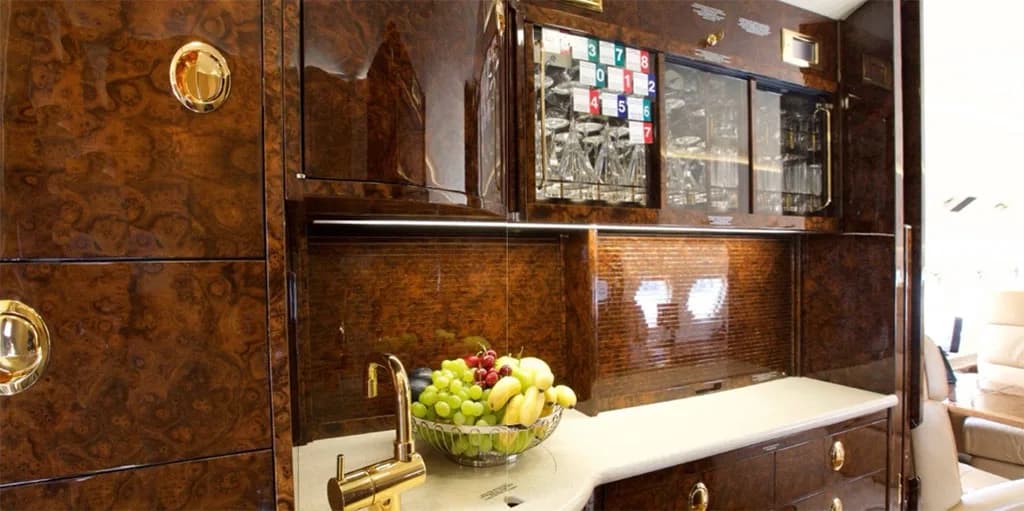 A close-up of the private jet's galley with dark wood cabinetry, a sink, and a bowl of fresh fruit on the counter.