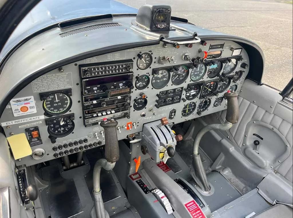 The cockpit of a small aircraft shows the instrument panel with gauges, radios, and control sticks.