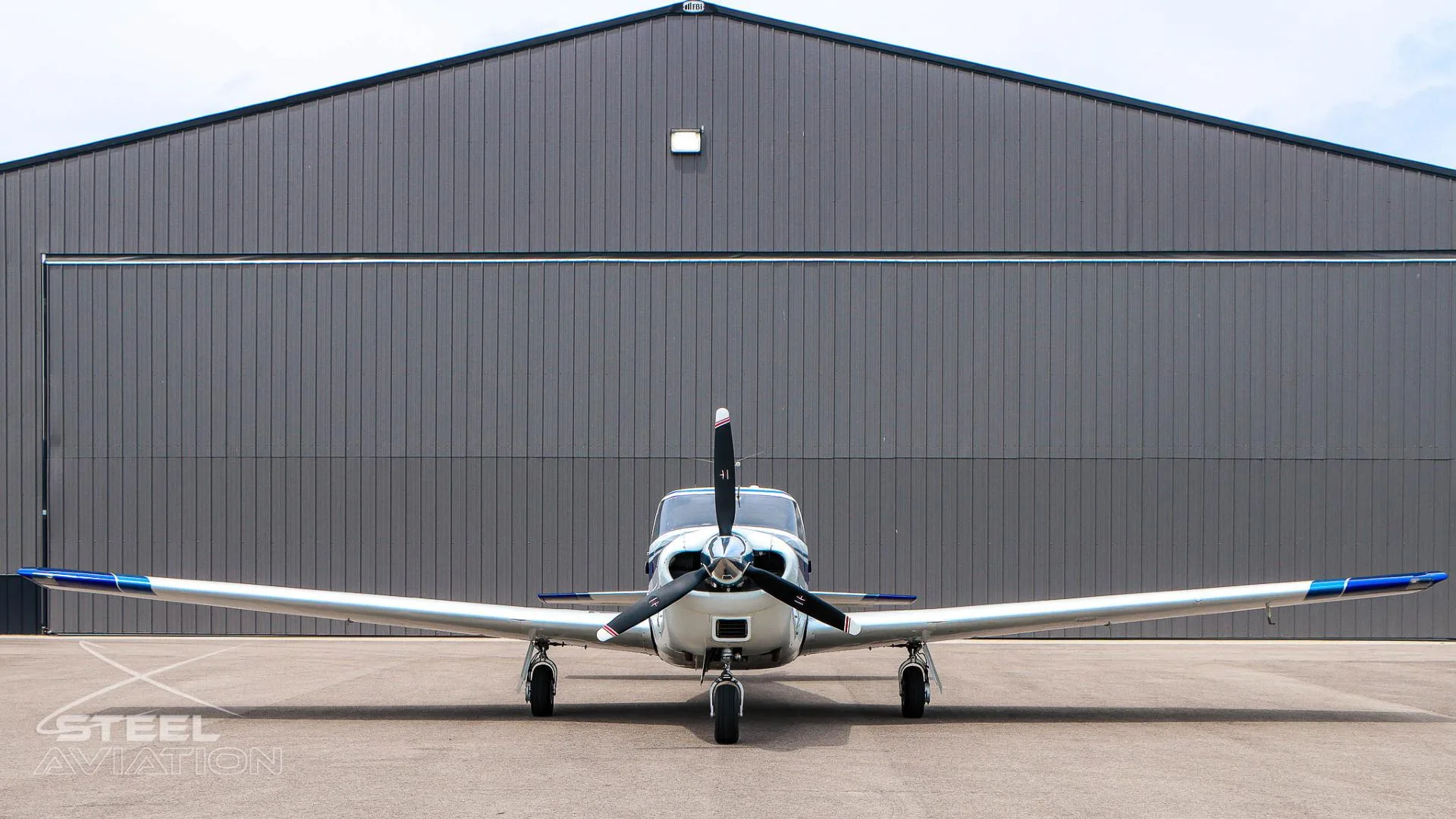 A head-on view of a white and blue single-engine aircraft parked in front of a dark gray hangar.