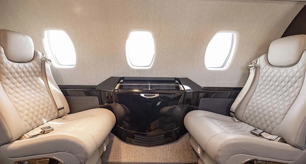 Two light gray quilted leather seats facing each other in the private jet's cabin, with dark wood cabinetry between them.