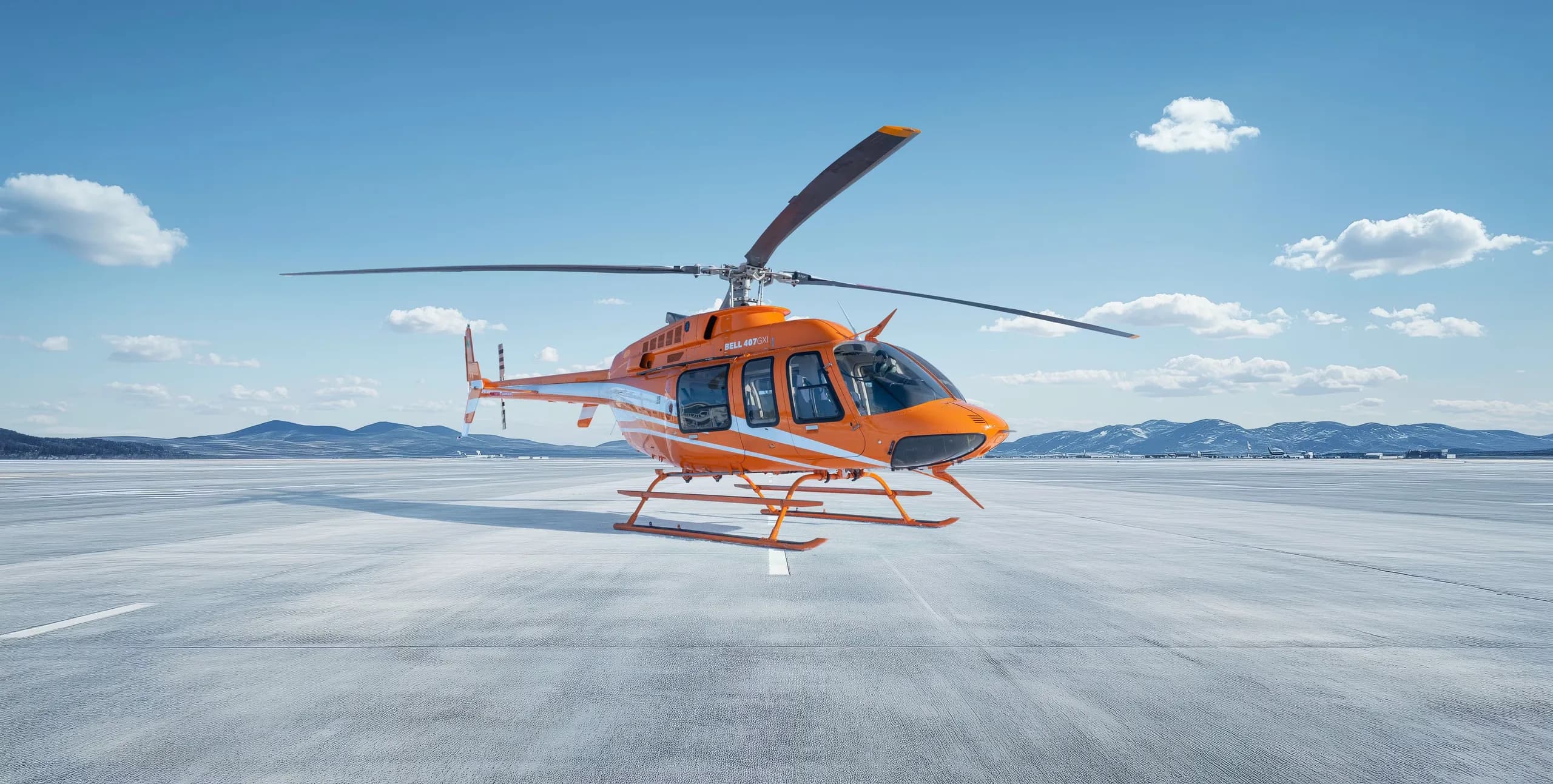 A Bell 407 helicopter with an orange and white paint scheme is parked on a concrete surface.