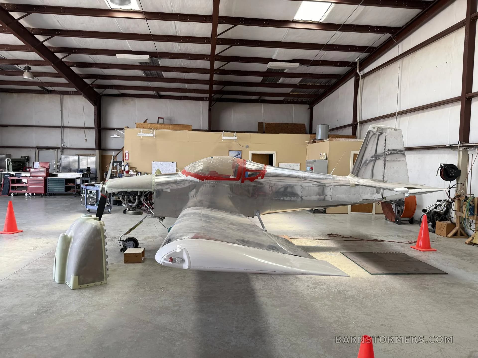 A side view of a partially assembled, unpainted metal aircraft with a red-taped canopy in a hangar.