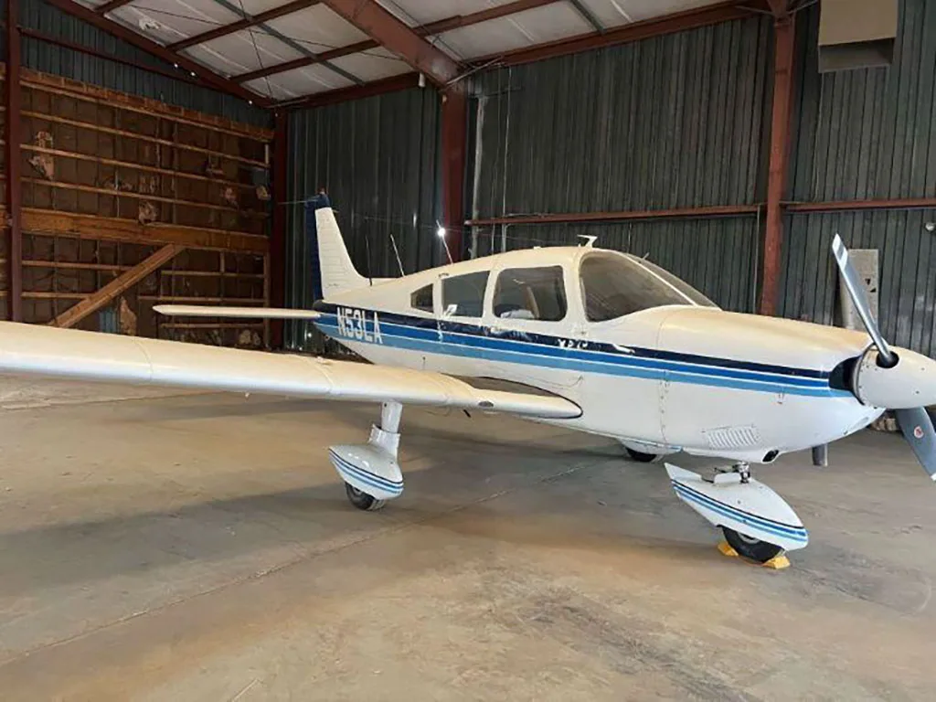 A front-right view of the white Piper Cherokee aircraft N53LA with blue stripes, parked in a hangar.