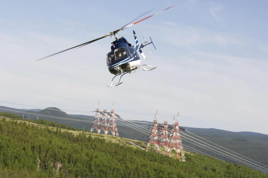 Bell 407 helicopter flying over a mountainous landscape with power lines.
