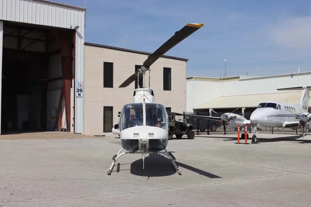 Front view of a white Bell 206 helicopter with its main rotor blade angled upwards, parked on an airfield.