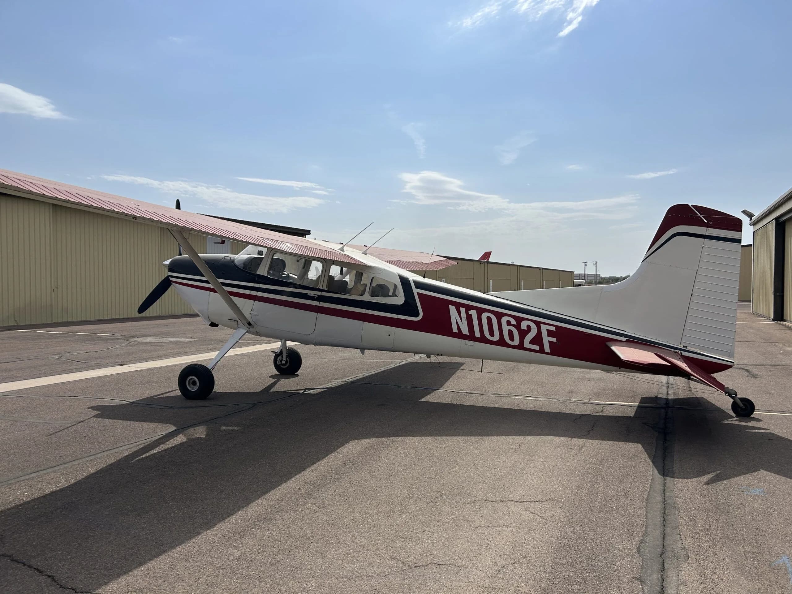 A white, black, and red Cessna 180 aircraft (N1062F) parked on an airport tarmac, viewed from the front-left.