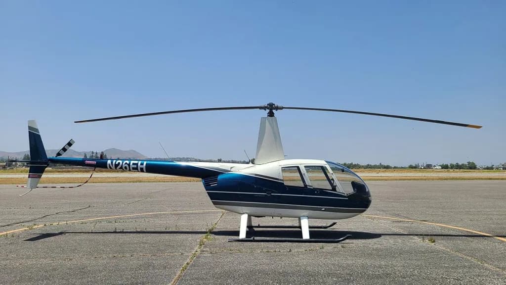 A blue and white Robinson R44 helicopter, registration N26EH, is parked on an asphalt tarmac under a clear sky.