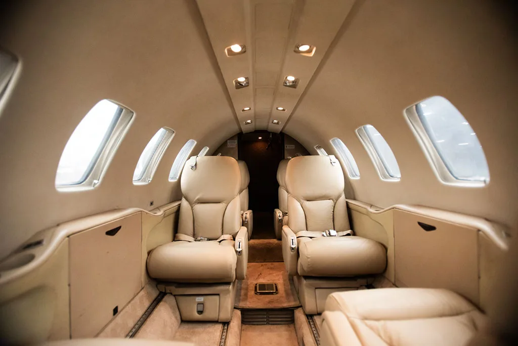 A private jet's beige leather cabin interior with two forward-facing seats and overhead lighting.
