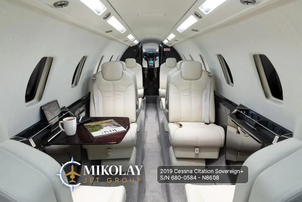 View of the aircraft cabin interior with light-colored leather seats, a table with a coffee cup and magazine, looking towards the front.