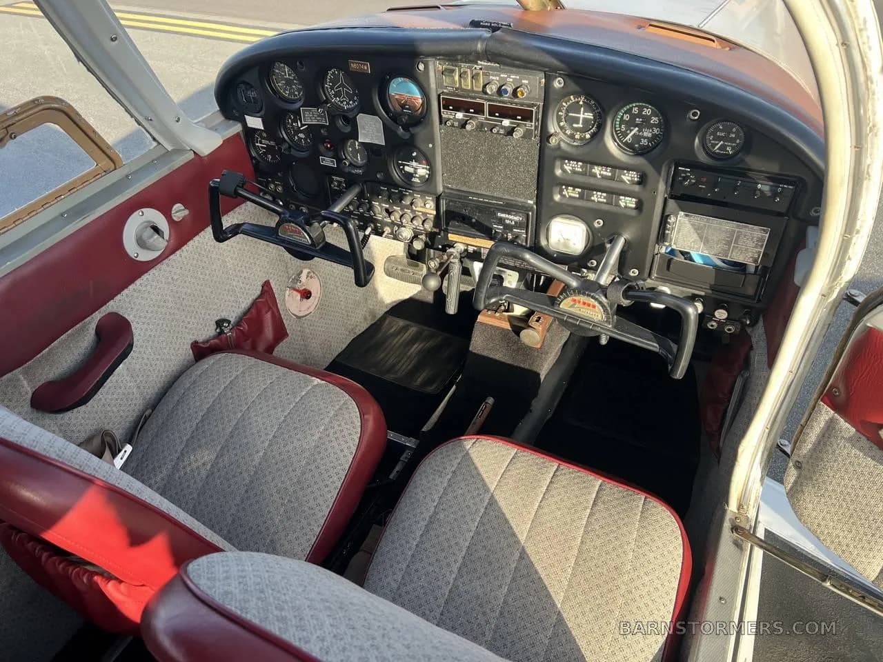 The cockpit of a small aircraft, showing the instrument panel, controls, and two front seats with red and patterned fabric.