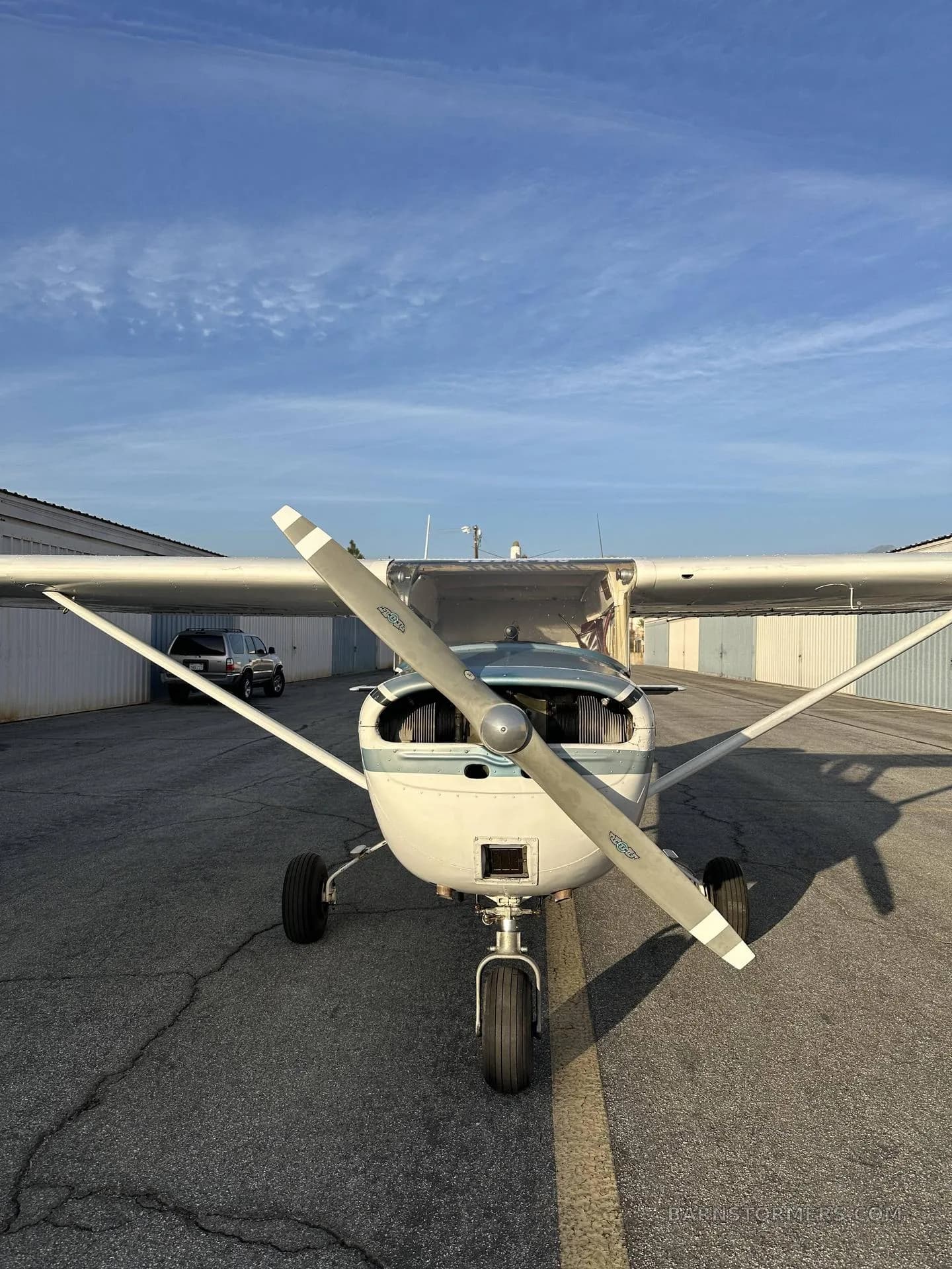 A front view of a white and blue Cessna 150/152 aircraft with its propeller centered, parked on a tarmac.