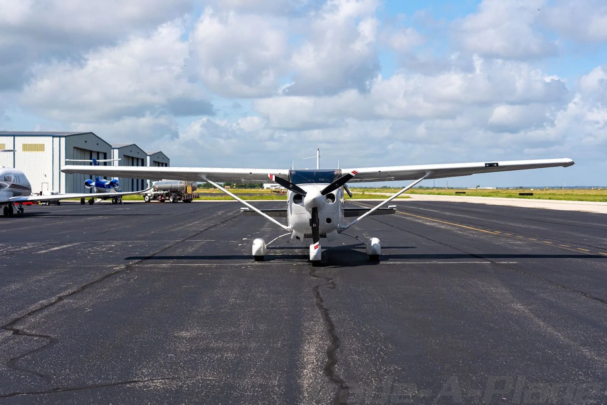 A front view of a white Cessna Stationair TC on a tarmac with hangars and other aircraft in the background.