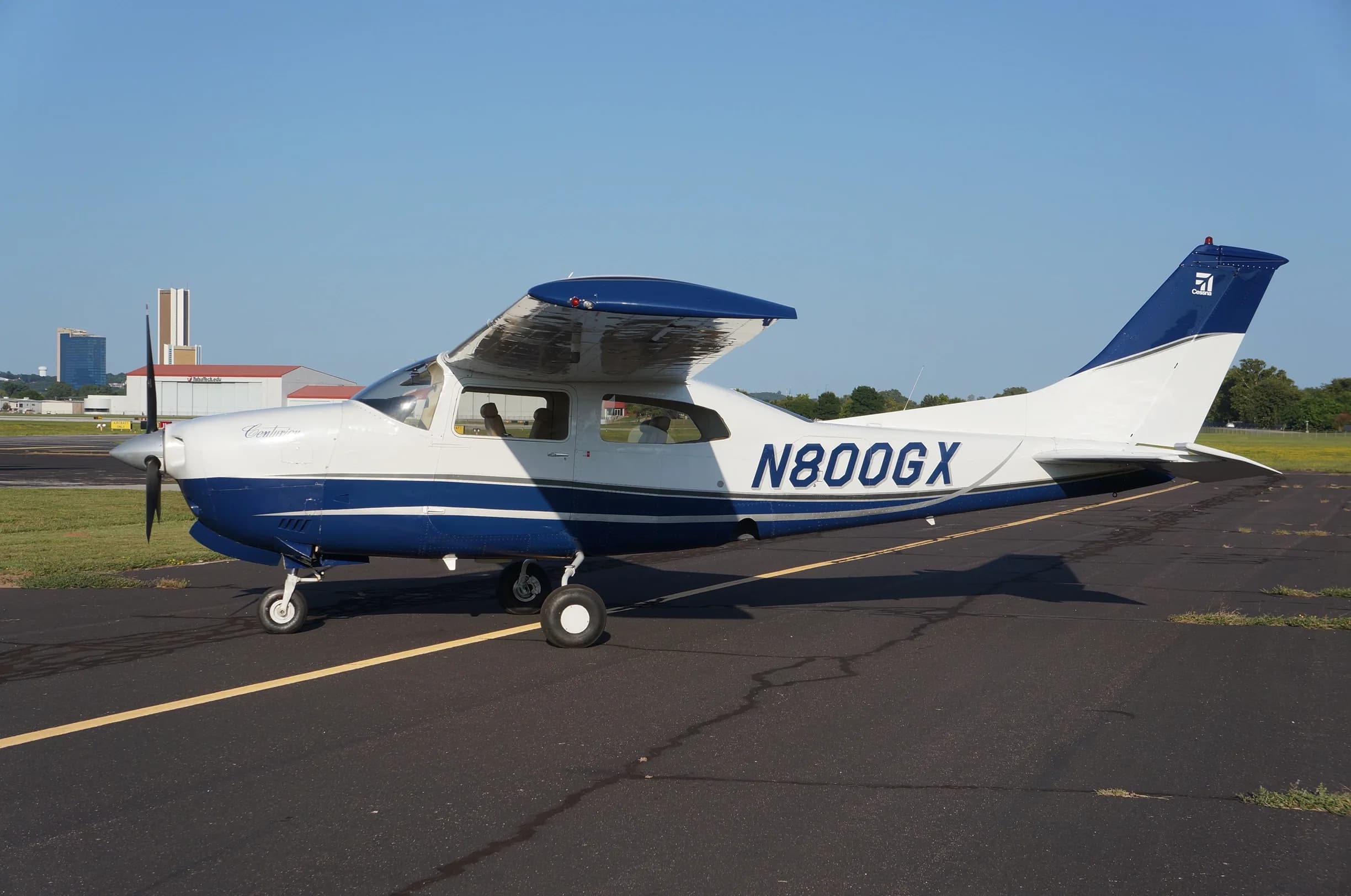 A white and blue Cessna 206 Stationair aircraft, N800GX, is parked on an airport tarmac with buildings and a city skyline in the background.