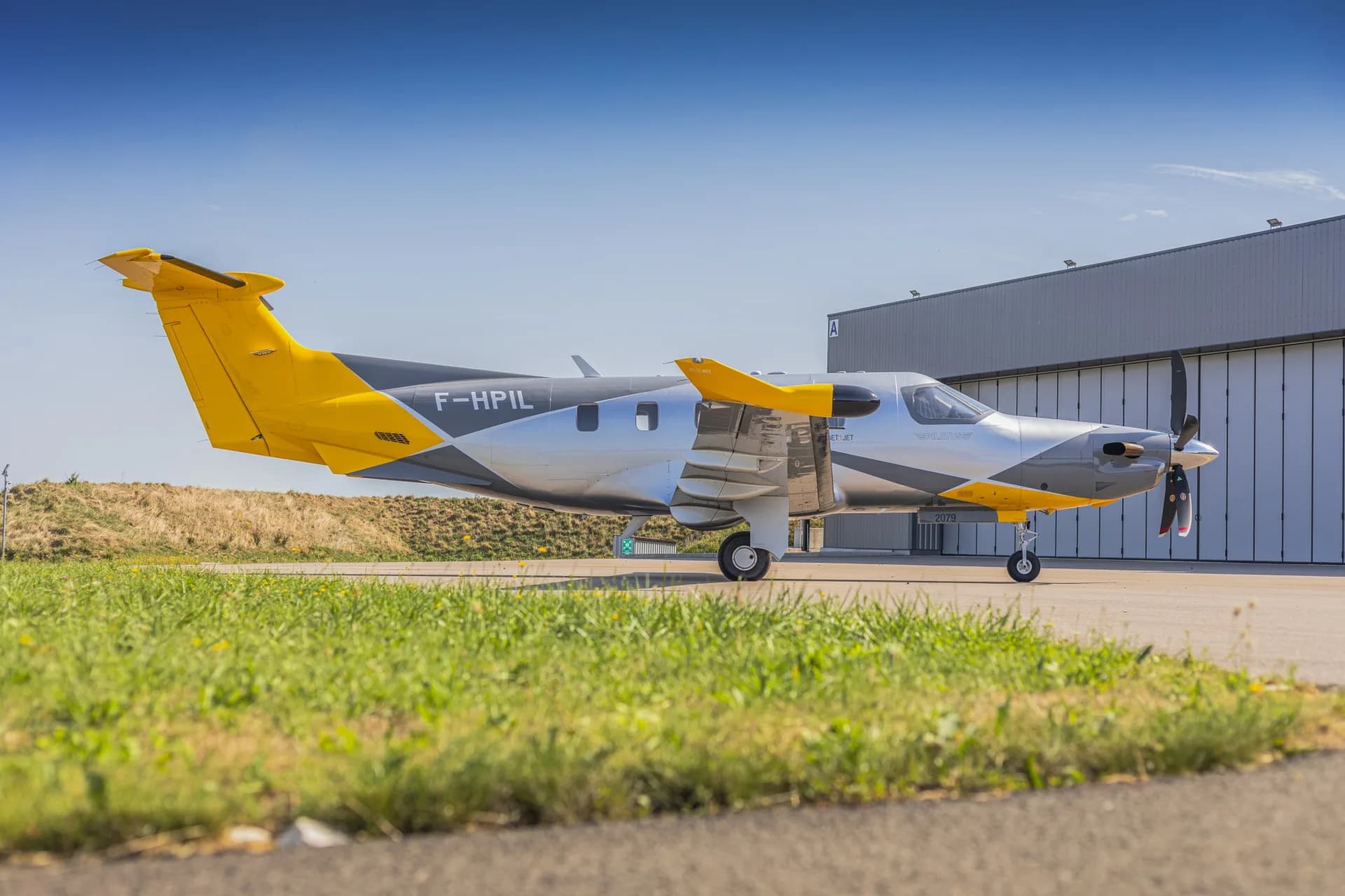 A side view of the silver and yellow Pilatus PC-12 aircraft (F-HPIL) parked on the tarmac near a hangar.