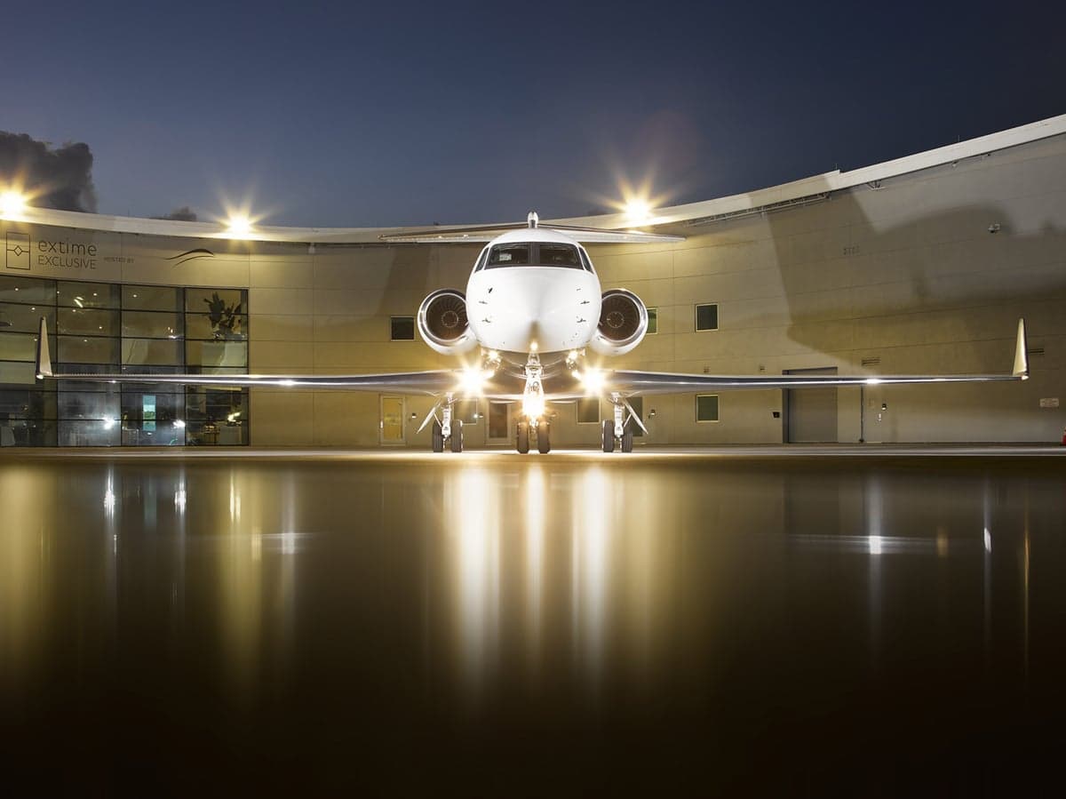 A white Gulfstream G650 business jet with its landing lights on, parked in front of a hangar at night.