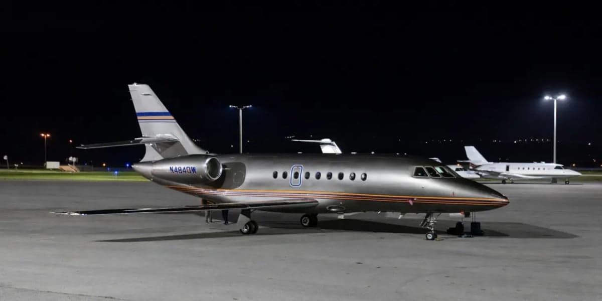 Side view of a silver Falcon 7X aircraft with orange accents on the tarmac at night.