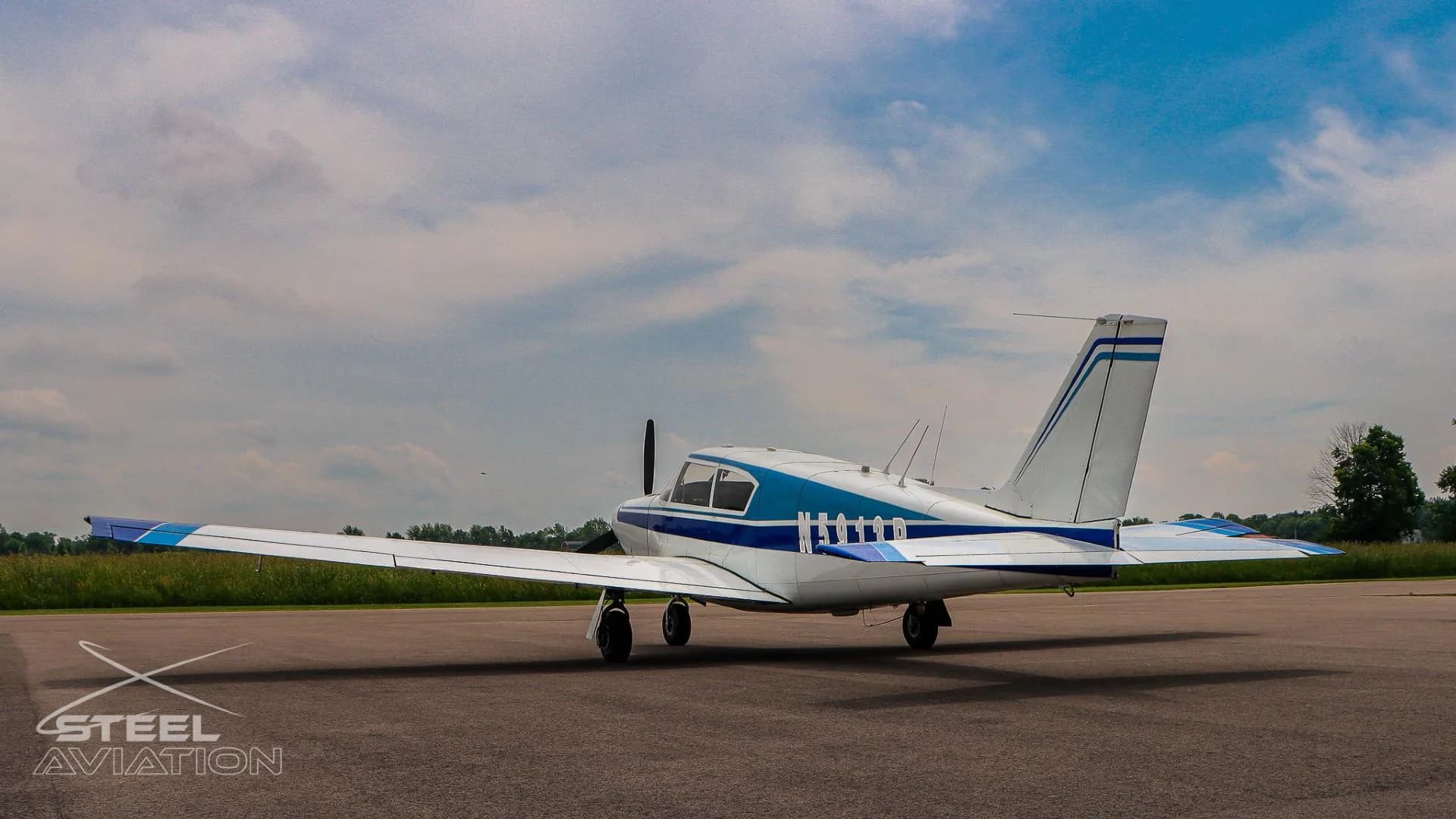 A rear three-quarter view of the white and blue single-engine aircraft N5913P on a tarmac with fields in the distance.