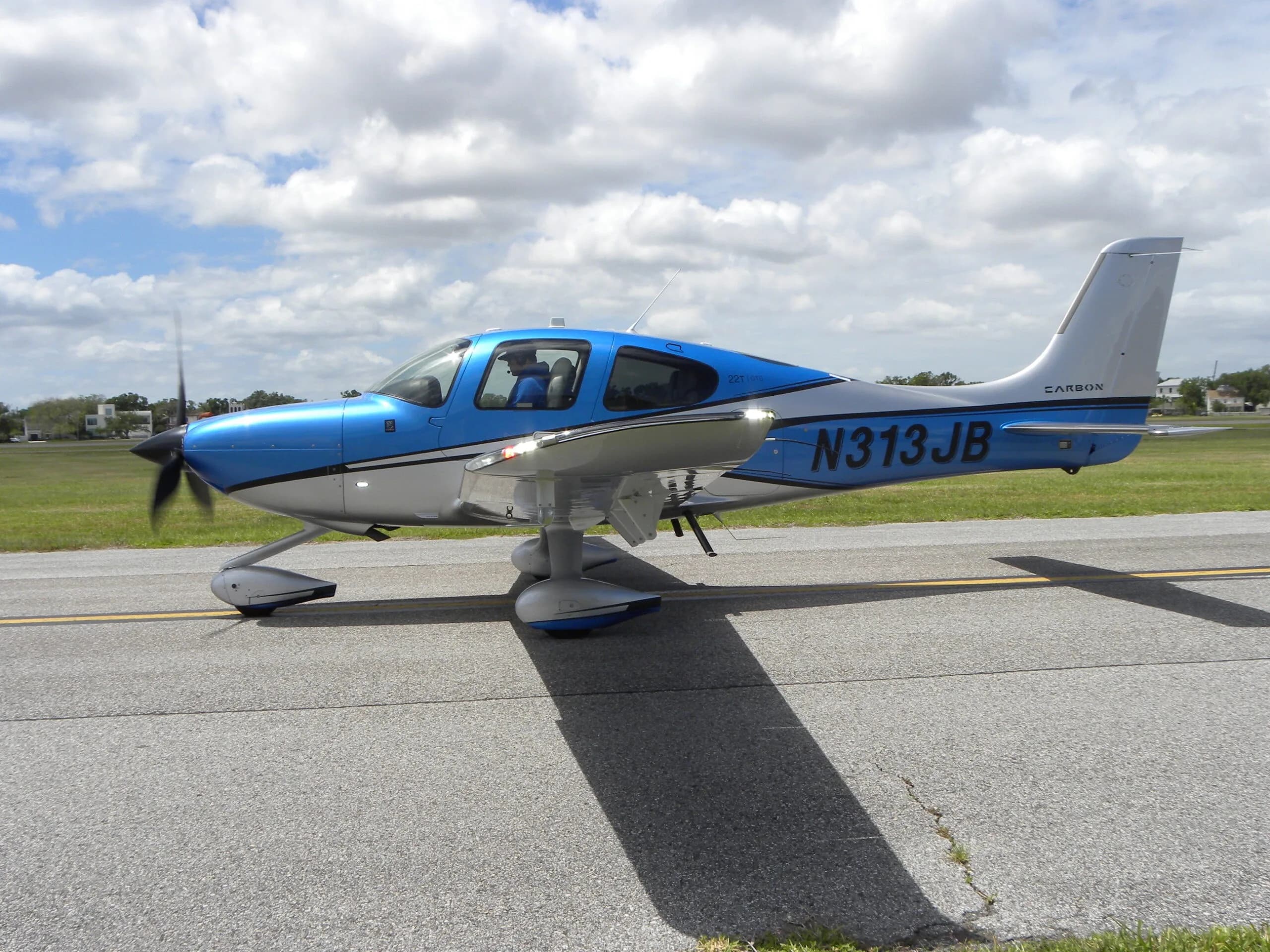 A blue and silver Cirrus SR22T aircraft, N313JB, with its propeller spinning on an airport taxiway.