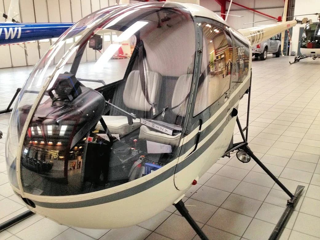 A white helicopter with grey stripes parked inside a hangar, showing its clear bubble canopy and interior.