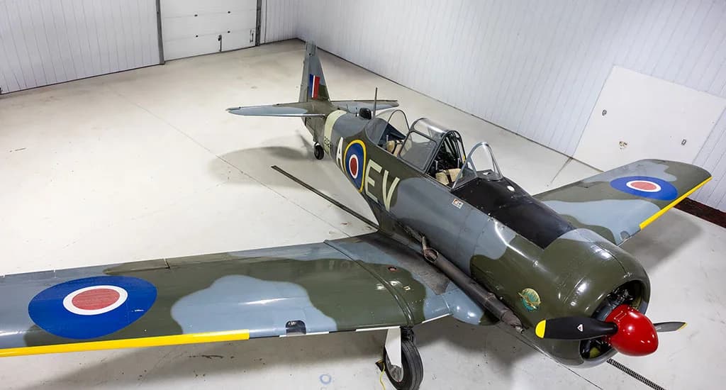 An overhead view of a North American Harvard Mk.IIB aircraft in green and grey camouflage with RAF roundels, parked in a hangar.