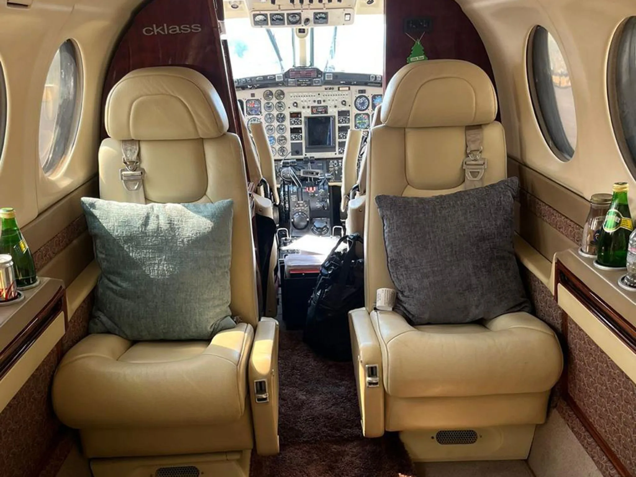 The aircraft cabin interior with two forward-facing light leather seats, each with a grey pillow, looking into the cockpit.