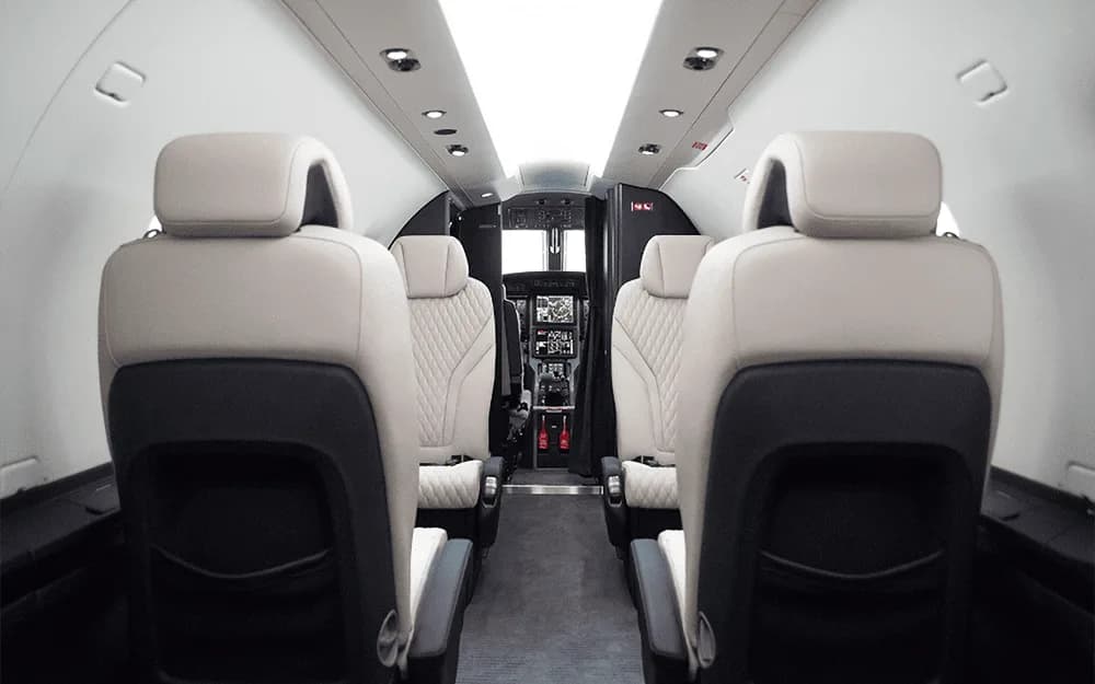 A view down the aisle of the Pilatus PC-12 NGX cabin, showing the white and black quilted leather seats and the cockpit beyond.