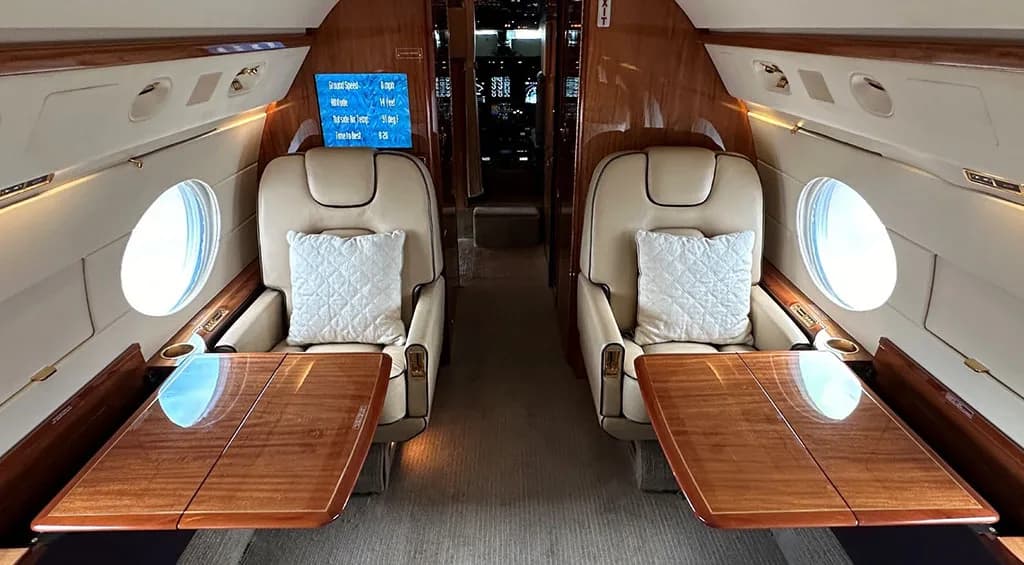 The luxurious interior of a private jet cabin with two facing beige leather seats and a wooden table.