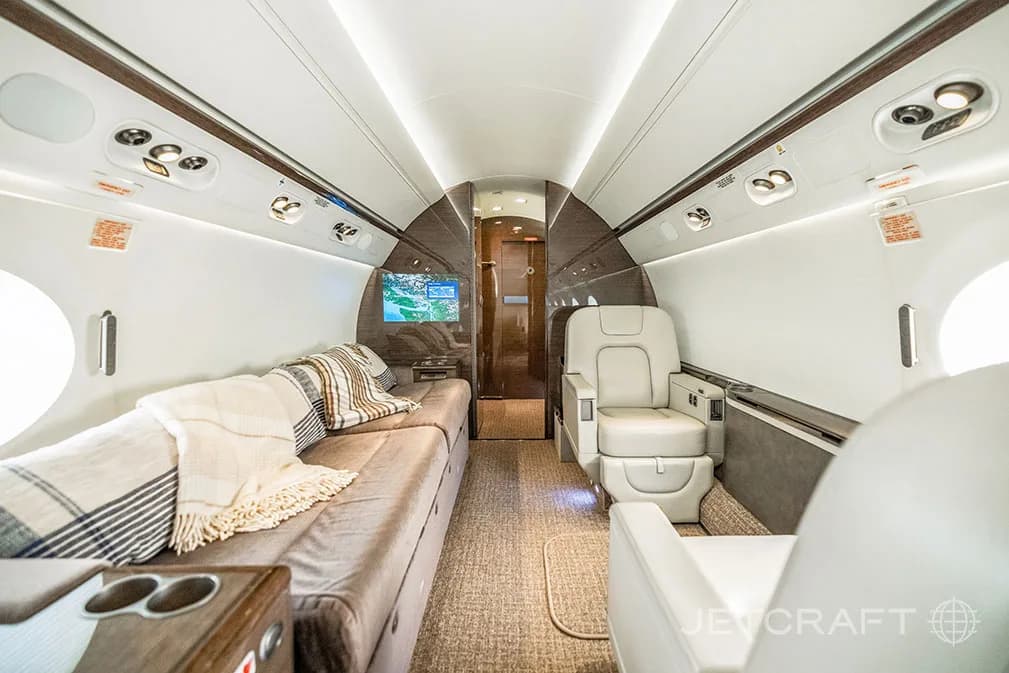 A private jet cabin with a brown sofa bed on the left, individual seats on the right, and a TV screen at the far end.
